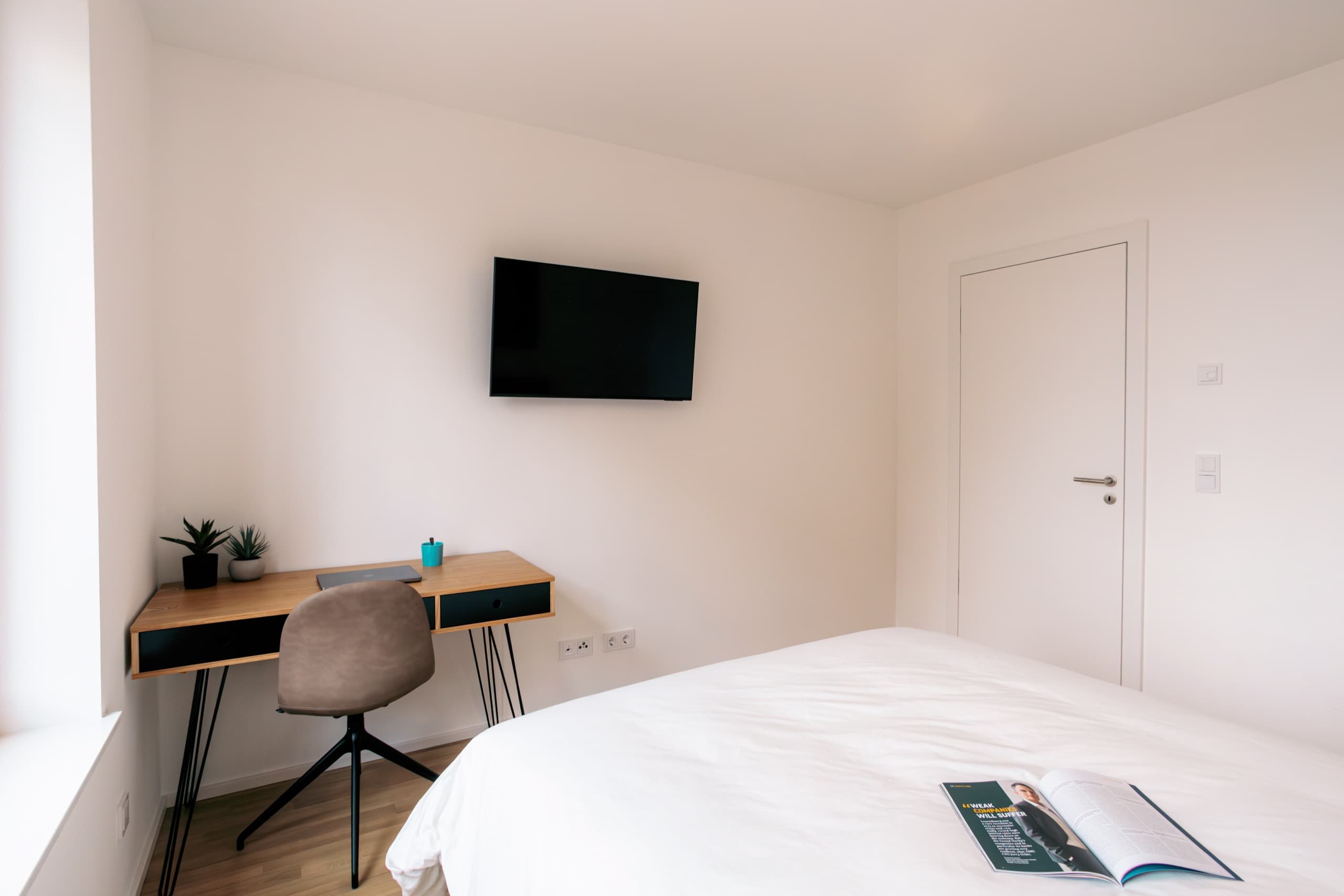 Bedroom in Apartment Escher at Media Bay, Kirchberg, Luxembourg.  There is a TV on the wall and a wooden desk in the corner.