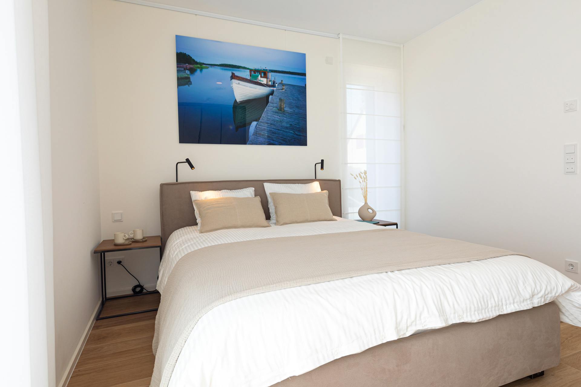 Bedroom with a large bed and white sheets and cushions, flanked by two lamps and nightstands. Hanging on the wall is a big picture with a fishing boat on water.