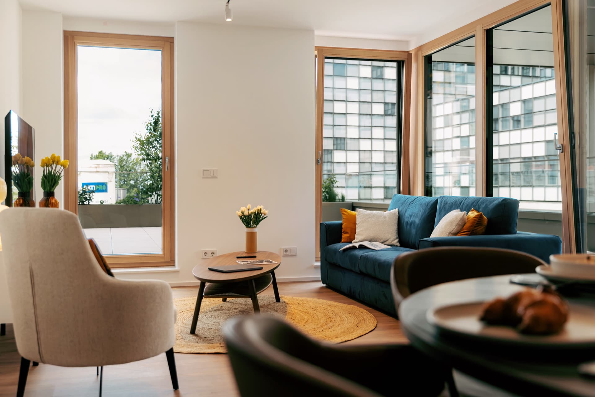 Picture of the lounge at Apartment Linde in Media Bay Apartments.  There is a soft blue sofa with white and gold cushions and in front a wooden coffee table with flowers sitting in a vase.  through the large windows behind the sofa you can see a very large terrace and the RTL office building behind. 