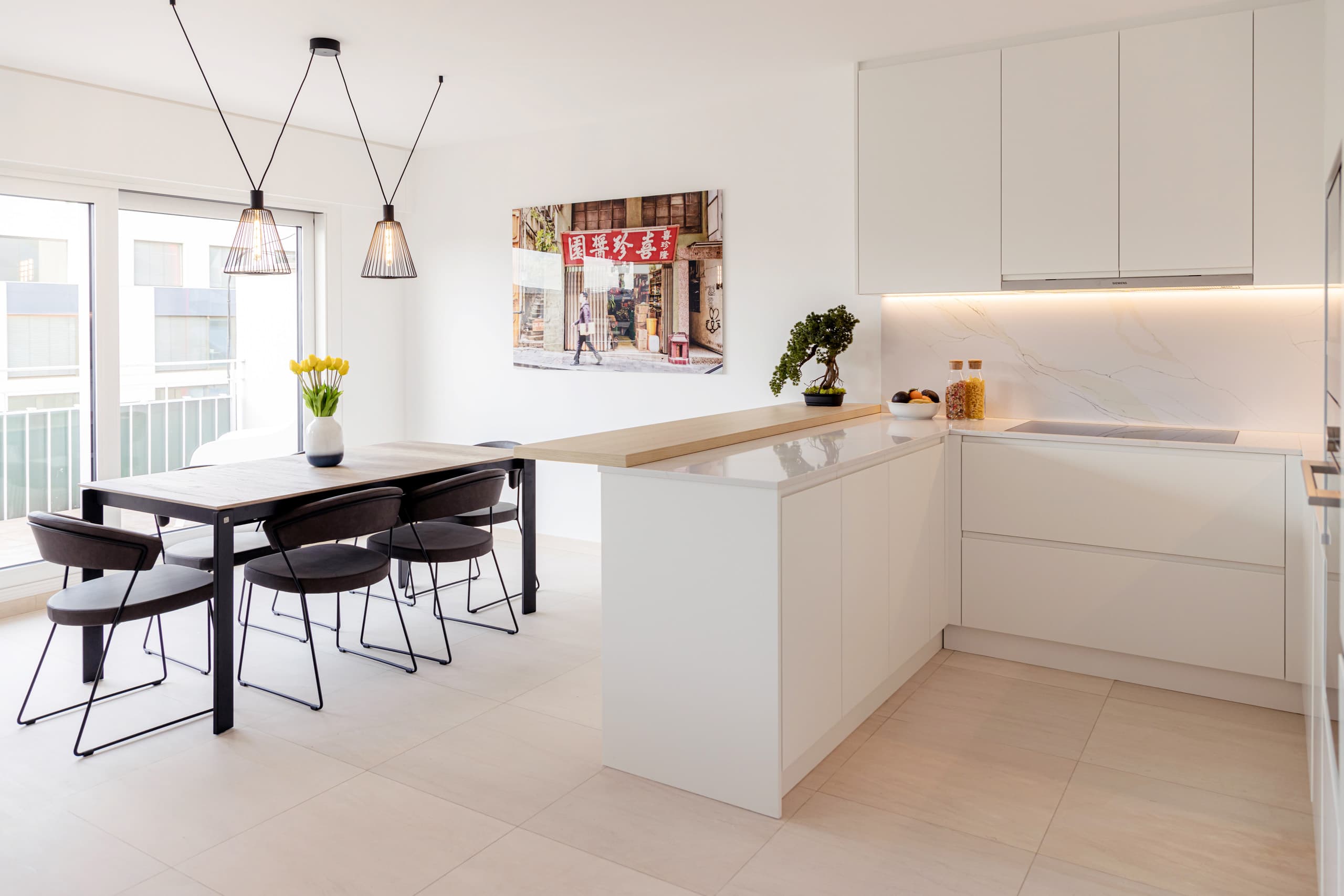 Penthouse for short to mid-term rent in Luxembourg city center, Hamilius area. The image depicts an interior view featuring a kitchen and dining area with a vase with yellow tulips on the table, a photograph of Hong Kong on the wall, a bonsai tree, fruit, and bottles with dried food objects on the kitchen countertop.
