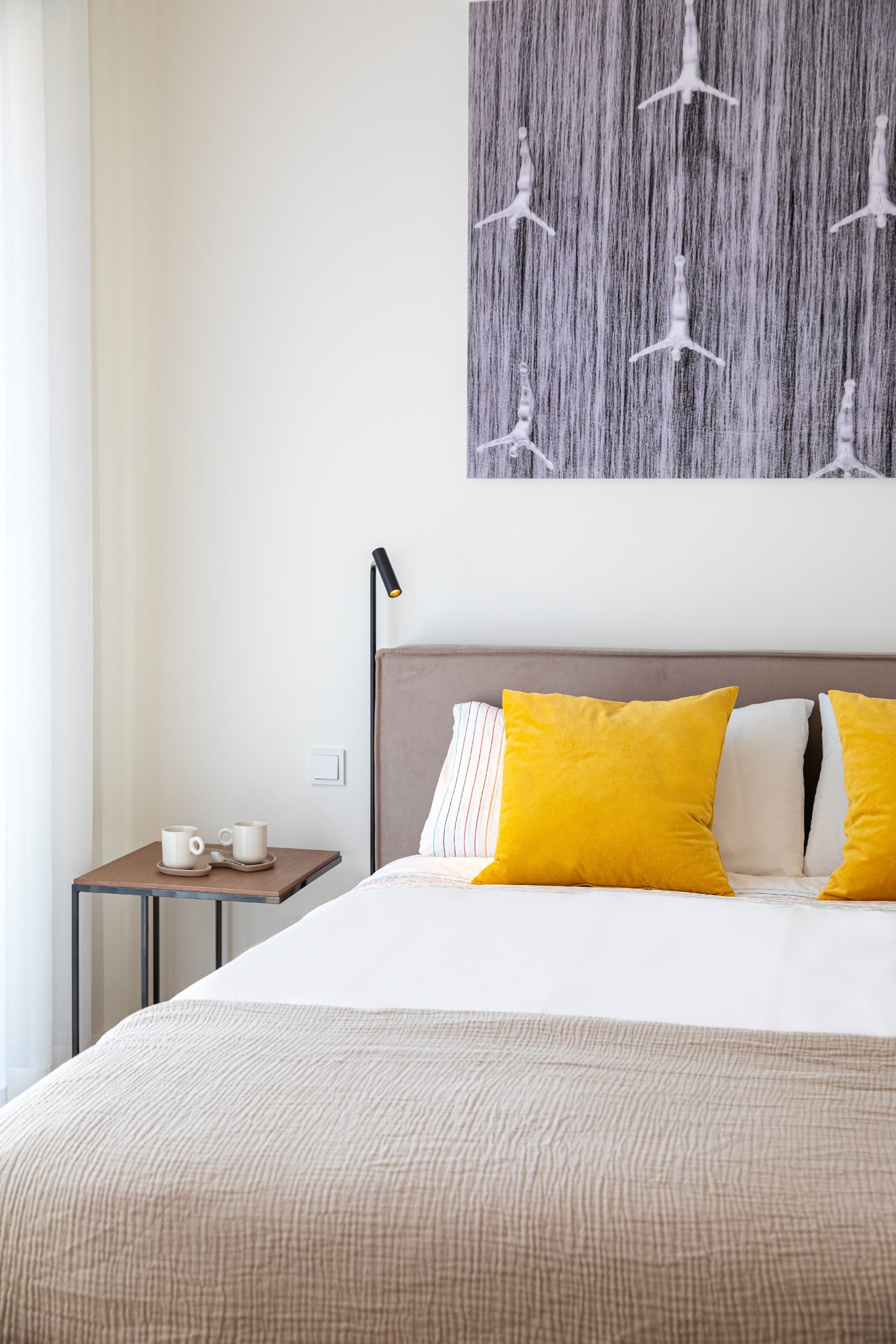 A bed in a bright room with white bedding and mustard coloured cushions. Next to the bed there is a side table with some coffee cups and a photograph of swimmers above.