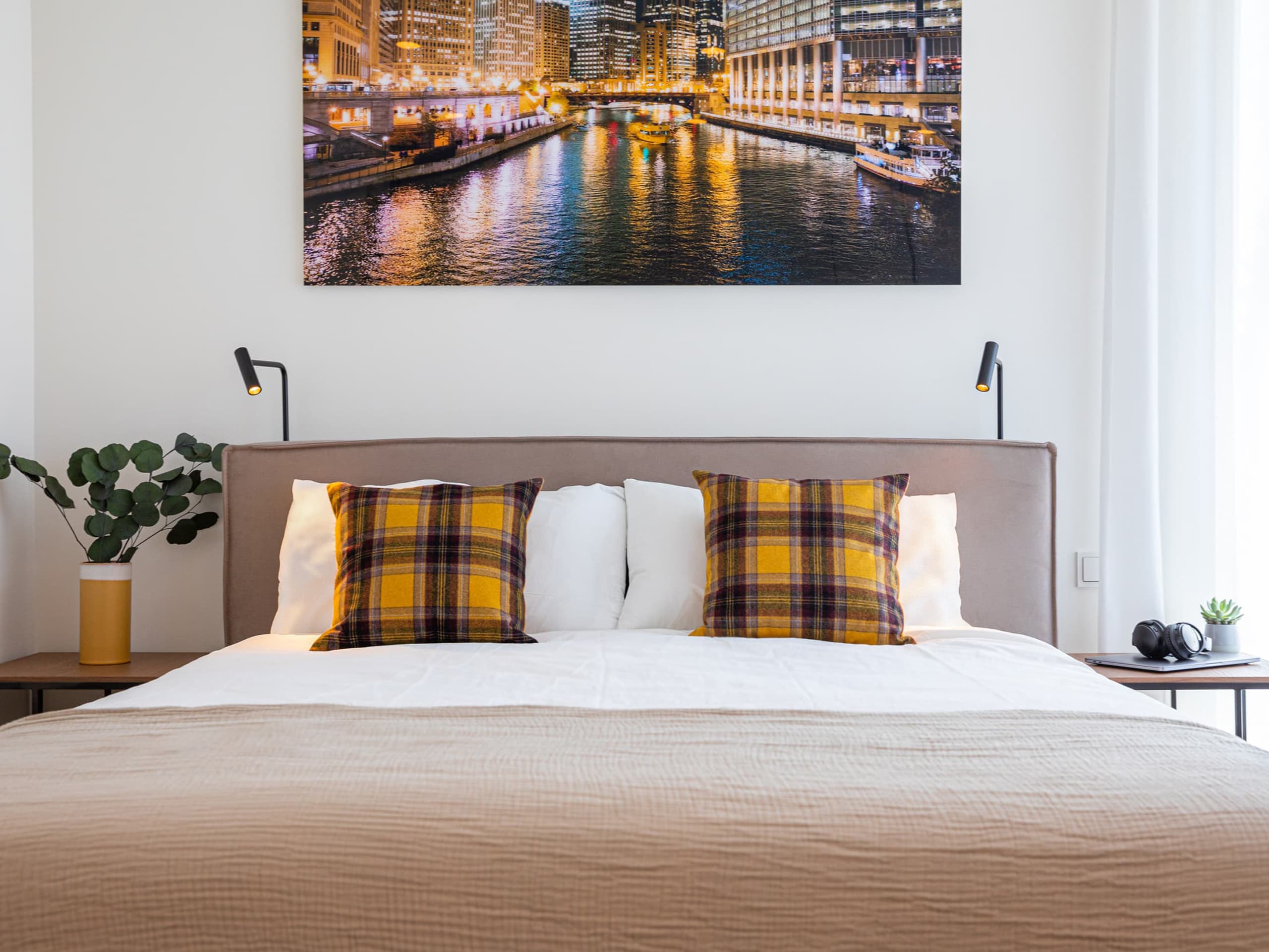 The picture depicts a bed with mustard tartan cushions and white bedding with a soft Alcantara headboard in beige. Above the bed, on the wall, there is a city view at night big photograph.
