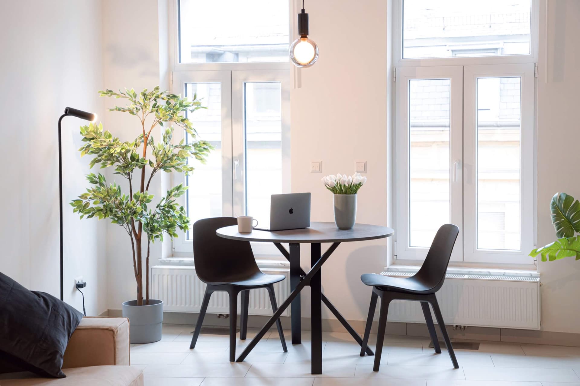 The inside of an apartment looking towards the 2 windows. In front there is a table with 2 chairs. Sitting on top of the table is a macbook pro, a vase with white tulips and a coffee cup. Next to the window is a large green plant.