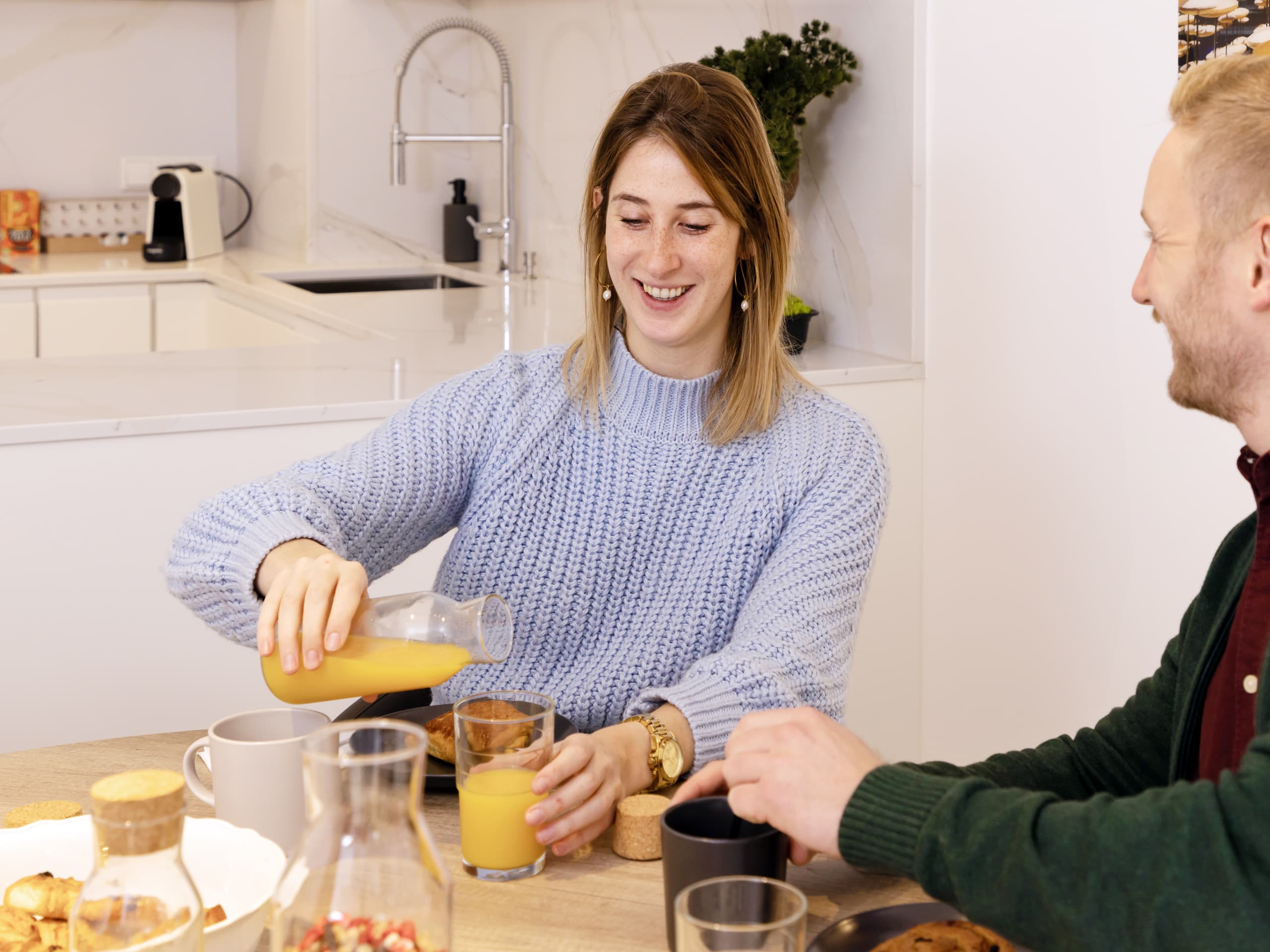 The picture shows a man and a woman having breakfast, the woman pouring orange juice at a dining table. In the background, you can see a white fully-equipped kitchen.