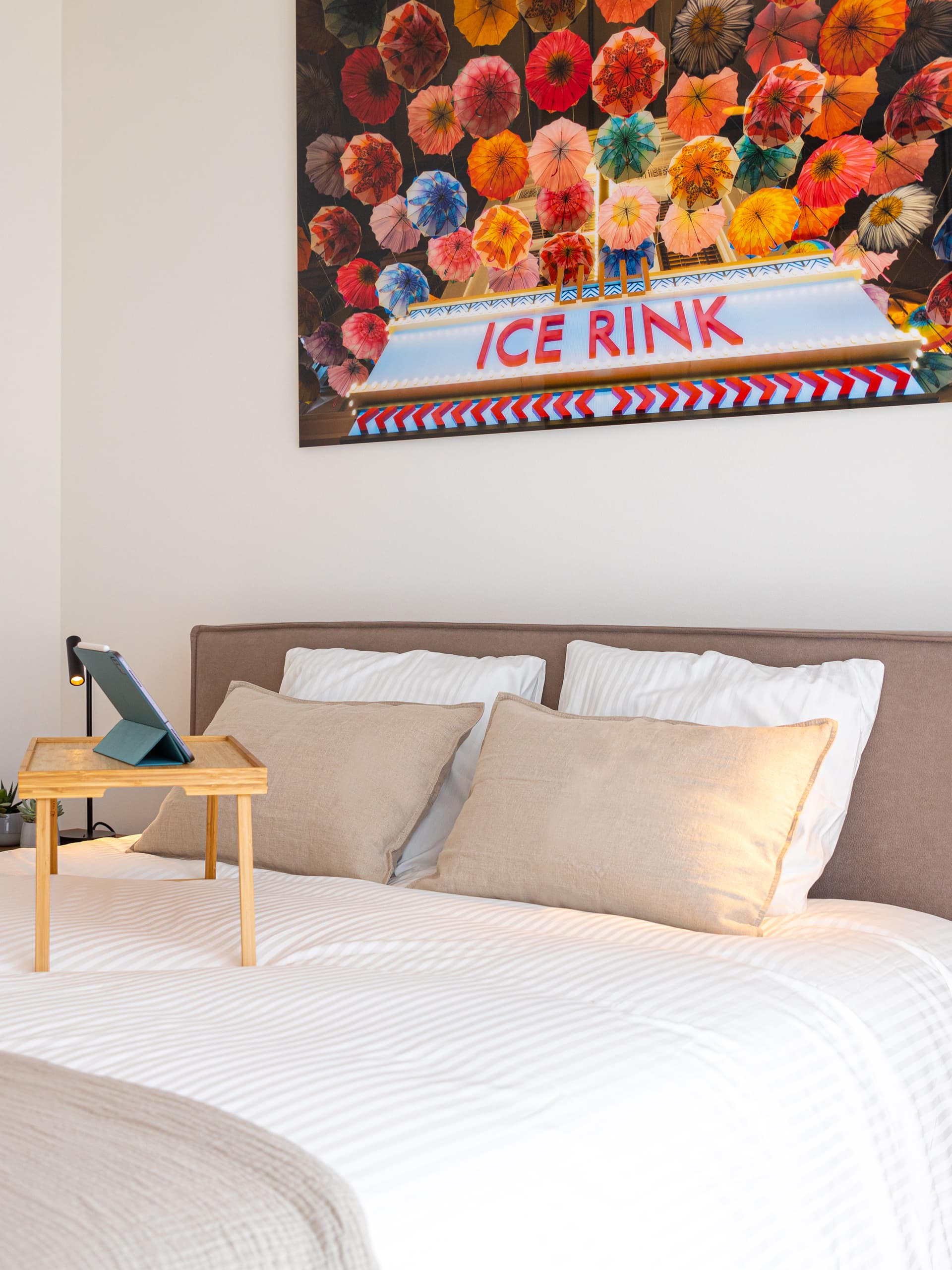 The picture illustrates the view of a bed with an iPad on a wooden-legged stand tray. Above the bed frame is a photograph of flowers and the words Ice Rink.