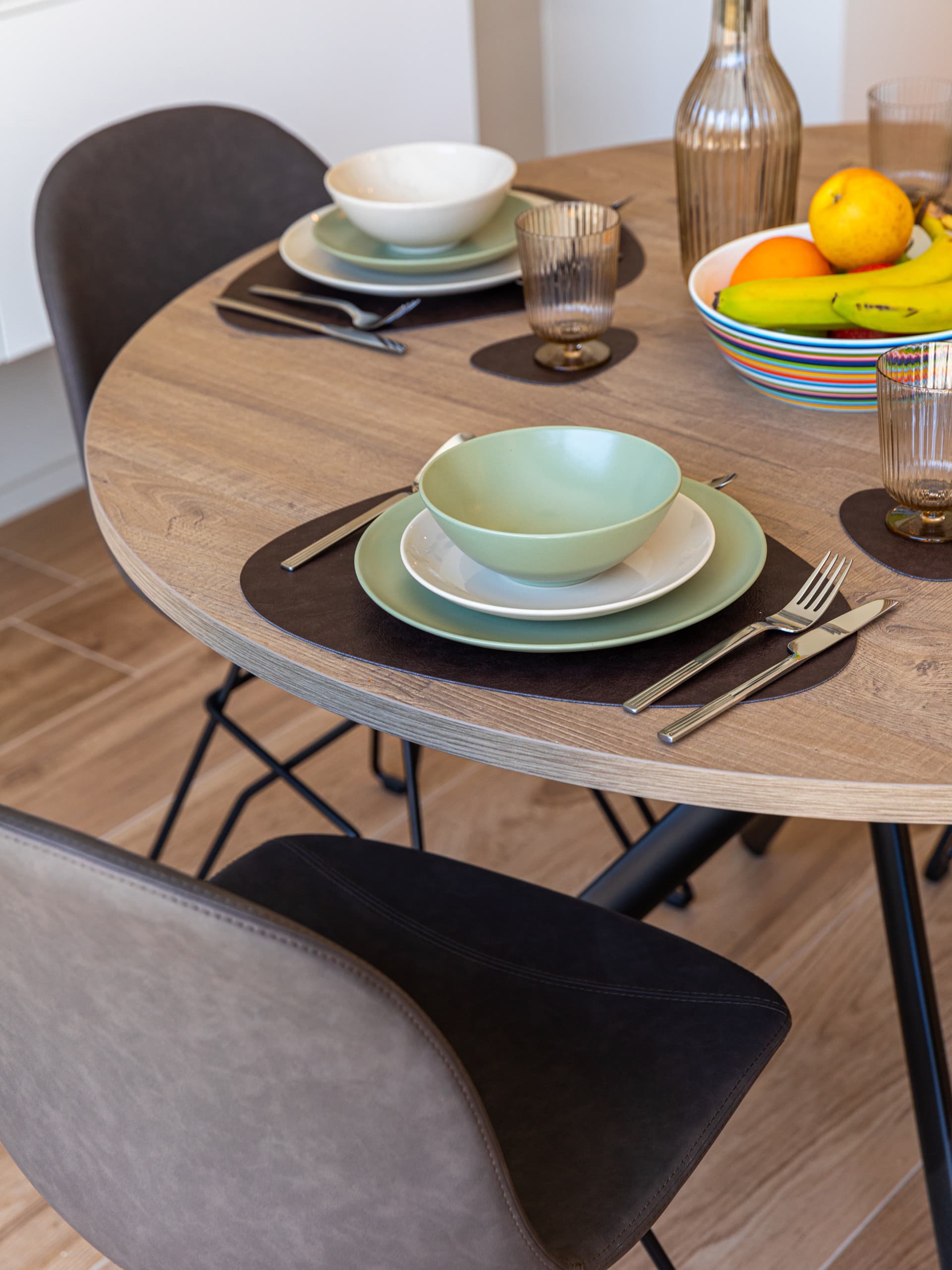 The picture depicts a dining table with several plates, glasses, cutlery, and bowls set for dinner. In the center of the table, there is a bowl filled with fruits.