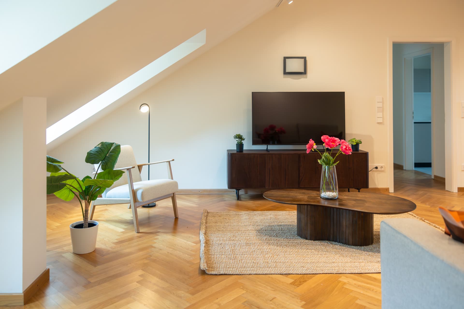 Lounge area with an armchair and a standing lamp next to it. In front, there is a mahogany coffee table with a flower vase on top. On the left, a TV stand with a large flat screen TV on it. A beige carpet over wooden parquet lights up the colors of the room.