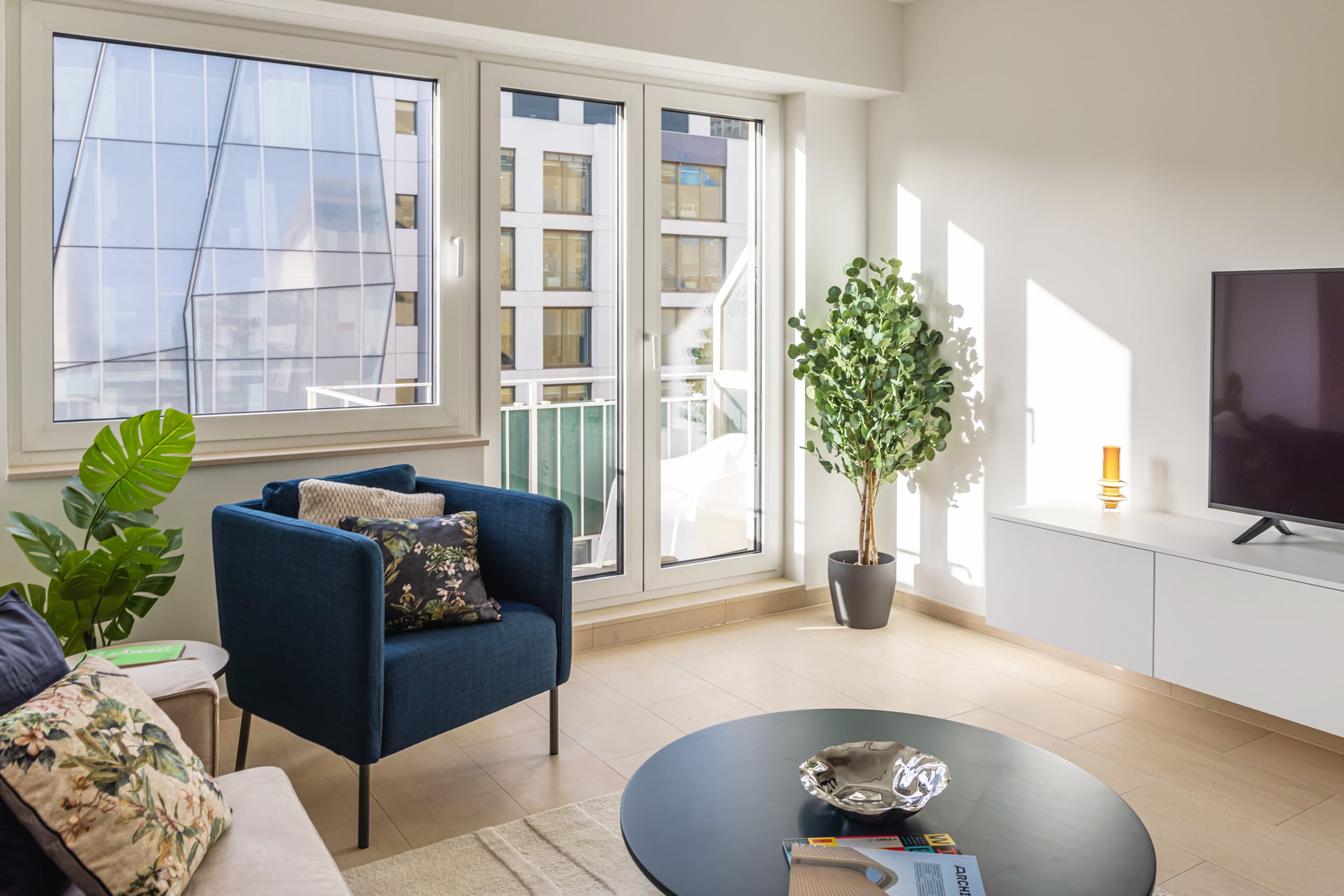 Seating area with a blue armchair and beige cushions next to a balcony doors with a large plant next to the window.